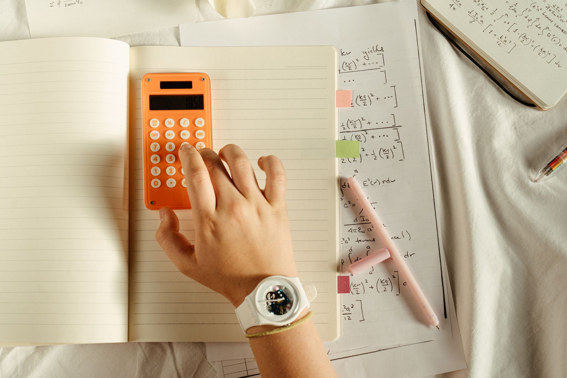 Nahaufnahme von Händen, die einen Taschenrechner und einen Notizblock auf einem leeren Tisch halten.
